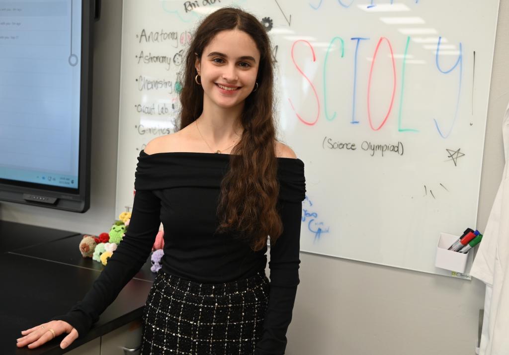 A young woman with long brown hair smiles in a classroom with a whiteboard that reads 'SCIOLY! (Science Olympiad)'.